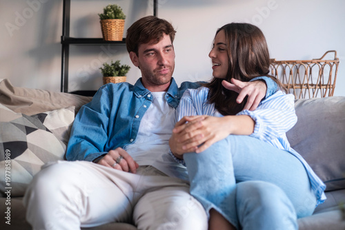 Tableau sur toile A young couple sits close on a sofa, smiling and talking while relaxing in a bright, cozy living room