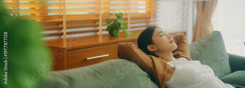 Relaxed young Asian woman enjoying rest on comfortable sofa, calm attractive girl relaxing on couch, breathing fresh air with eyes closed, meditating at home
