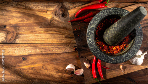 Top-down view of a stone mortar and pestle filled with crushed red chili surrounded by fresh peppers and garlic cloves on a dark wooden rustic surface with dramatic shadows
