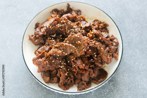 Top view of marinated stir-fried beef slices, sprinkled with white sesame seeds. The dish is presented on a white plate set on a dark grey background.