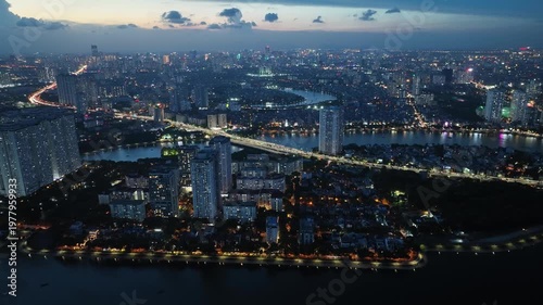 Twilight aerial view of Linh Dam Lake with glowing city lights, dense urban skyline, and soft evening sky transitioning into night