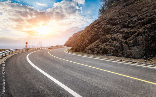 Winding asphalt mountain road with safety guardrail at sunset. Scenic curvy highway during golden hour