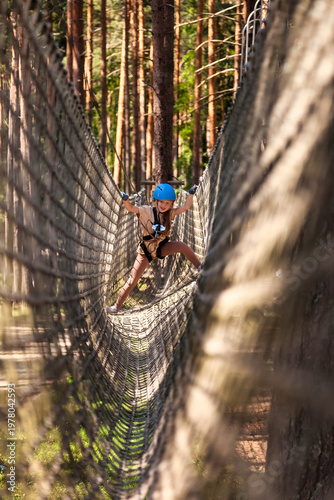 A young girl navigates a challenging rope course suspended between trees on a sunny day. The vibrant greenery surrounding her highlights the thrill of outdoor adventure and exploration.