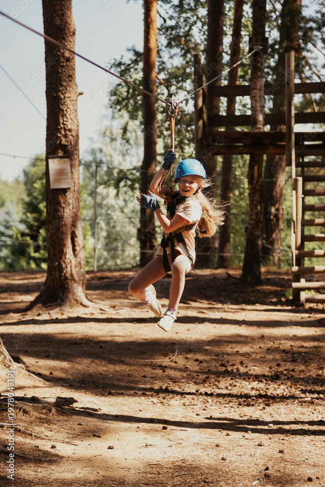 Fototapeta premium A girl is soaring through the air on a zip line, her face radiating joy and excitement. Dressed in a helmet and gloves, she is surrounded by tall trees and the adventurous spirit of the rope park.