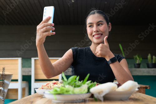 Happy Thai Woman Giving Thumbs Up Taking Selfie with Smartphone at Restaurant with Thai Food