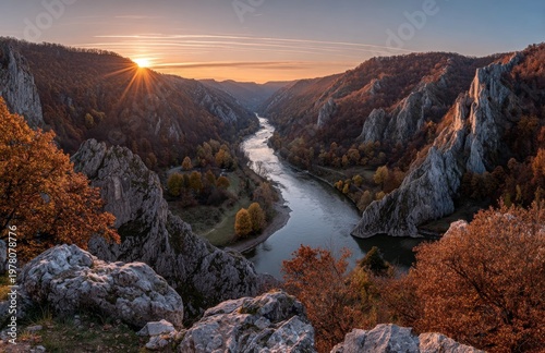 A golden sunrise bathes a mountain valley with a winding river, while the orange sky and clouds mirror on the water; green trees cloak the hillsides, and the rocky foreground is dotted with dry grass