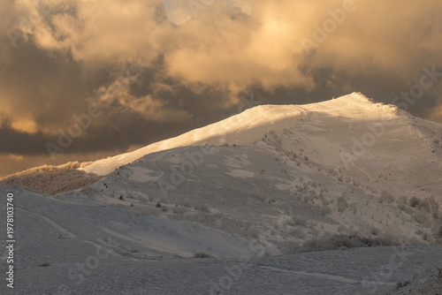 The majestic snow-covered peak of Smerek in the Bieszczady Mountains illuminated by bright sunlight against a backdrop of dramatic dark storm clouds.