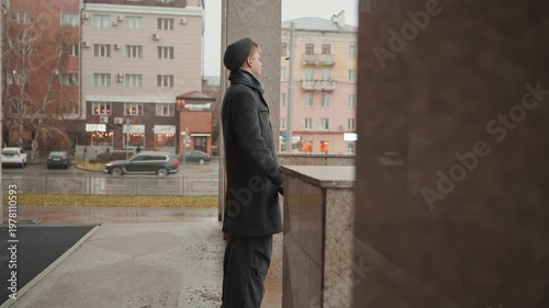 Thoughtful figure pauses amid bustling streets. Contemplative individual stands on wet stairs watching traffic move. Serene observer with collar up stands still as cars pass by silently