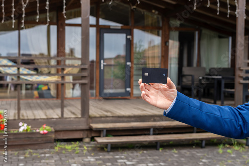 Plastic credit card in hand with new house at background. Broker, mortgage, property, real estate concept. Buying or renting