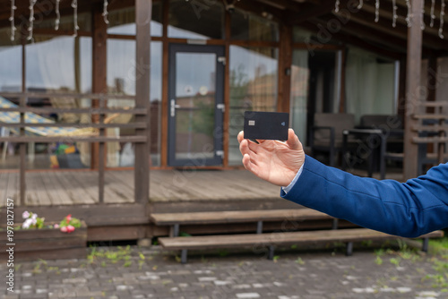 Plastic credit card in hand with new house at background. Broker, mortgage, property, real estate concept. Buying or renting