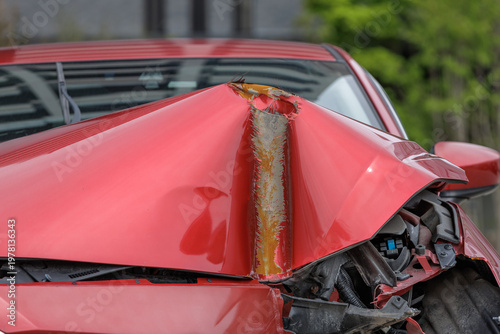 Fotografie Bright red car with deep vertical indentation from strong frontal impact shows severe front-end damage with hood crushed upward to reveal engine components and partially expose tire well