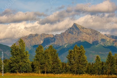 Scenic view of Krivan, one of the most iconic peaks of the High Tatras in Slovakia. Mountain ridge rises above green forest and meadow under partly cloudy sky, illuminated by warm natural light.