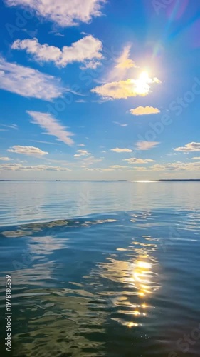 Sun shining over calm blue ocean water with fluffy clouds in the sky, peaceful seascape with bright sunlight reflecting on the sea surface.