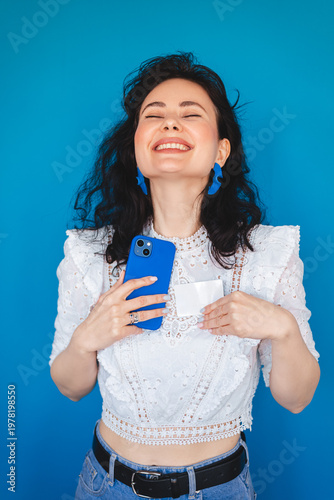 Extremely happy young woman laughing with closed eyes holding smartphone and credit card to her chest feeling relieved and joyful after successful purchase or big sale isolated on blue background