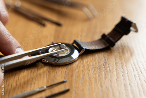 Quadro su tela Hand holding tweezers replaces a watch battery on a wooden surface, with watch t