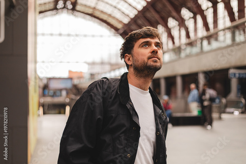 Thoughtful young man traveler looking up at information board or architecture while waiting for train departure standing in large modern railway station terminal with blurred historical background