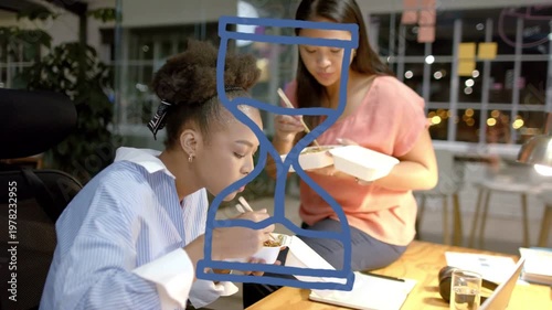 Two female coworkers in office working late, eating takeout, checking laptop while hourglass hiding