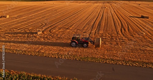 Agricultural tractor collecting hay bales in field at sunset