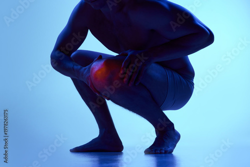 Canvas-taulu Man squatting holding knee with red pain highlight under blue neon lighting