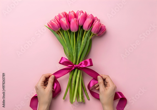 Hands tying a fuchsia ribbon around a fresh bouquet of pink tulips on a pastel pink background, top view
