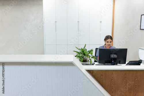African woman receptionist in grey scrubs using monitor behind wood counter with plant, copy space