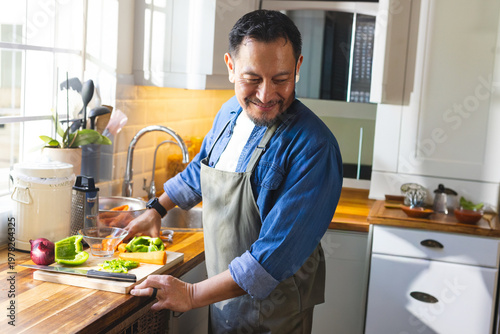 Mature Asian man in apron chopping pepper and red onion on wooden board in home kitchen