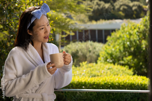 Asian woman standing on balcony holding beige mug wearing white bathrobe blue headband, copy space