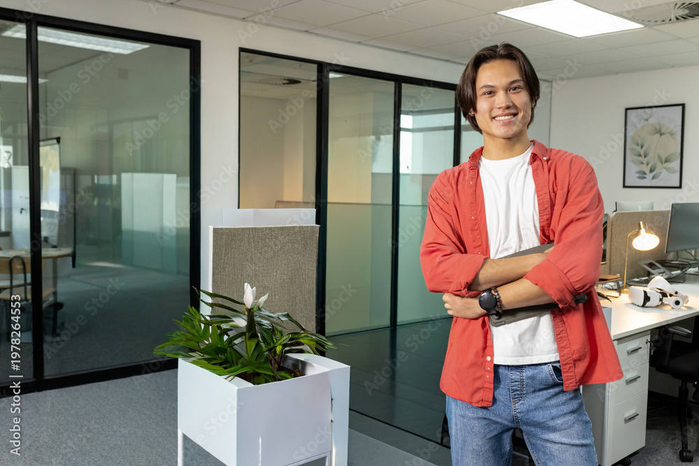 Fototapeta premium Asian man wearing red shirt and jeans standing in open-plan office near white planter, copy space