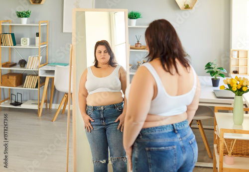 Obraz na płótnie Photo of obese fat woman dressed in white top and jeans, looking at her reflection in mirror, doesnt like her body, has access weight, gaining to be fit, needs to keep diet