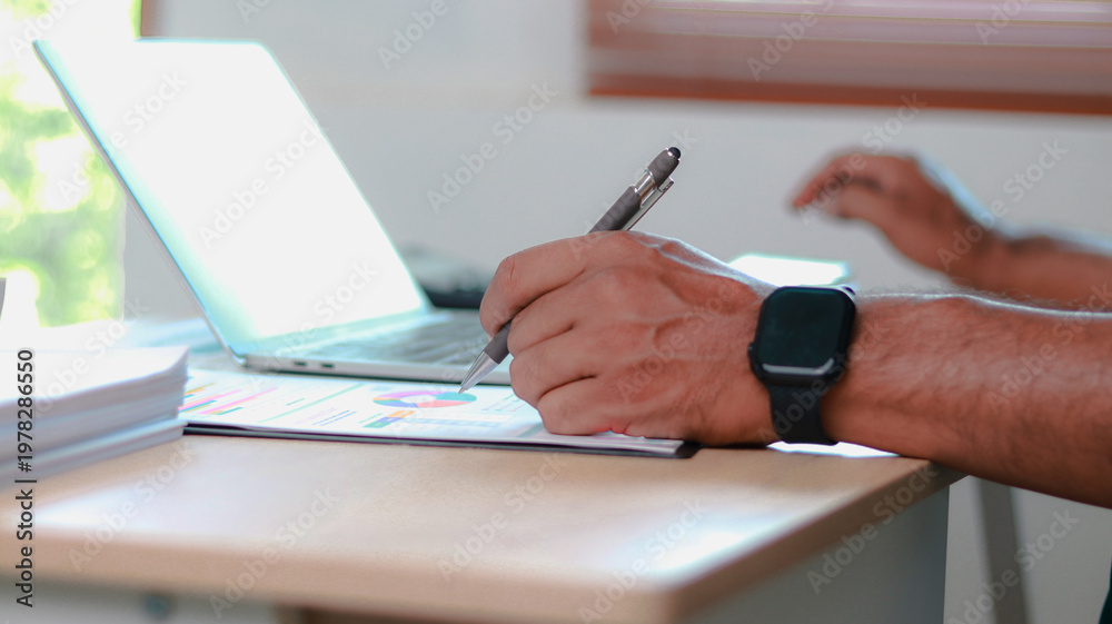 Fototapeta premium Close-up of professional analyzing business data, writing notes on report with pen while using laptop. Concept of data analysis, financial planning, productivity, and modern work.