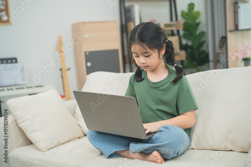Young girl studying on a laptop while sitting comfortably on a sofa in a cozy living room with plants and soft lighting