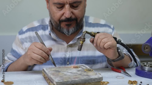 Wide shot of a jeweler working at a bench using a torch to shape and solder a gold jewelry piece. Shows full workspace, tools, and professional environment of jewelry production.