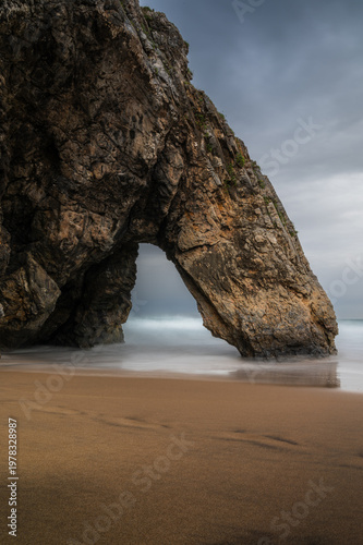 Cinematic long exposure shot of arch on Praia da Adraga beach on a moody day, Sintra, Portugal. Vertical travel banner