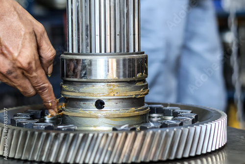 Canvas Print Technician applying grease to a heavy industrial metal shaft and bearing assembly in a workshop to provide lubrication during machinery maintenance