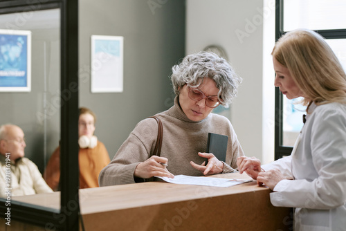Canvas Print Senior Caucasian woman signing medical form while female doctor guiding paperwork at clinic reception, representing patient intake and healthcare administration