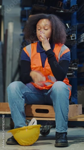 Fatigued African American female warehouse worker taking off a yellow hard hat and sitting down to rest after a hard workday.
