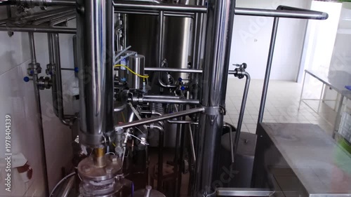 Processing room shows stainless tanks and pipework near walkway. Worker shadow crosses doorway shifting slowly behind valves and gauges