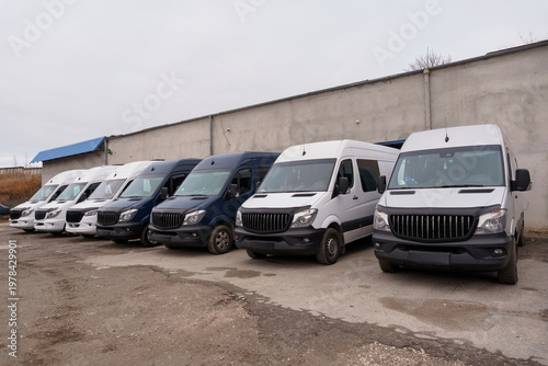 Fotografie Row of white and dark blue commercial delivery vans parked in a lot against a concrete wall on a cloudy day