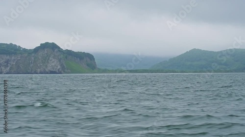 Waves against the backdrop of the mountainous coast of Avacha Bay in the Pacific Ocean of the Kamchatka Peninsula in cloudy, overcast weather