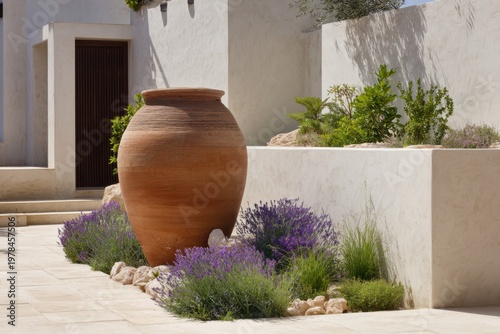A large clay pot stands next to purple flowers and green plants in a yard with white walls and steps under a clear blue sky during the day