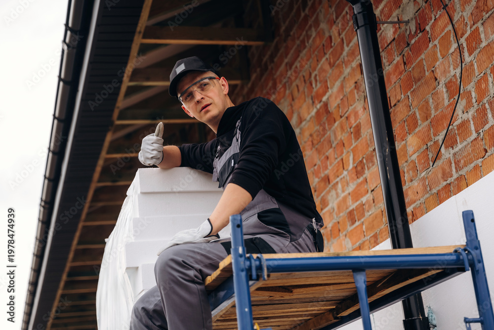 Fototapeta premium Young male construction worker in gray overalls and black cap gives a thumbs-up while sitting on scaffolding with stacked white materials against a brick wall