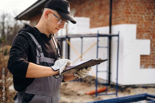Wallpaper Mural Male construction worker in gray overalls and cap reviews clipboard while standing near a building site with scaffolding and insulation materials in the background Torontodigital.ca