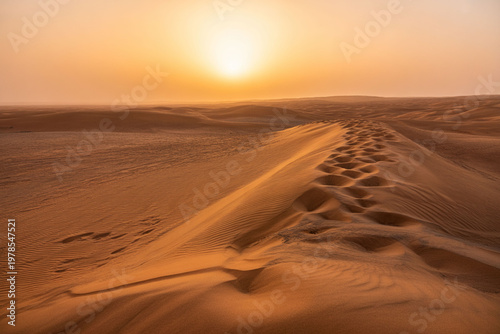Golden desert landscape at sunset with sand dunes footprints and warm hazy sky creating sense of vastness exploration.