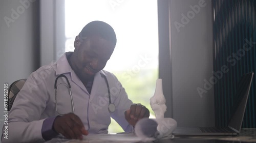 African American male doctor writing prescription on clipboard at desk, Orthopedic physician working with medical record, laptop and knee model in clinic, Healthcare and physical therapy concept
