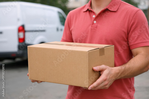 Delivery worker holding cardboard box, white van parked on street with blurred buildings in background