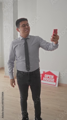Man holds red smartphone for selfie beside open house sign in empty building; property listing confidence.