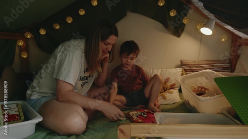 Mother and son engage in playful activities on a bed inside a cozy fort, surrounded by soft lighting and colorful toys