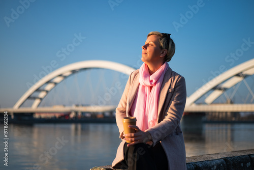 Portrait of relaxed woman enjoying a coffee by the river, eyes closed in sunlight, taking a peaceful break from busy city life.