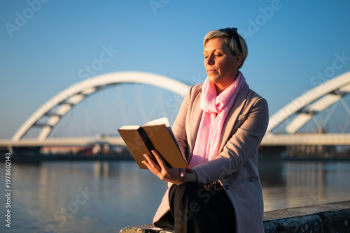 Portrait of modern woman with short blonde hair reading a book while enjoying sitting by the city river on a sunny day.