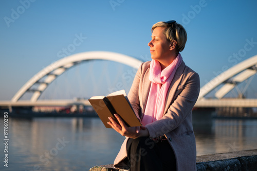 Portrait of beautiful relaxed woman reading book by the river in a city, capturing a peaceful lifestyle moment filled with joy, reflection and natural light.